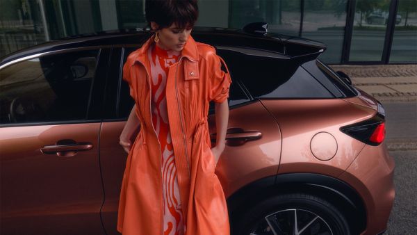 Eine Person in einem orangefarbenen Trenchcoat und einem gemusterten Kleid steht neben einem bronzenen Lexus LBX