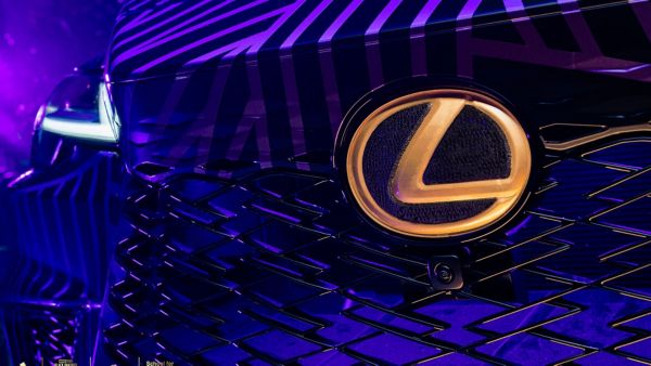 Nahaufnahme eines Lexus-Kühlergrills mit dem markanten Lexus-Logo in Gold vor einem lila Hintergrund mit auffälligen schwarzen Streifen.