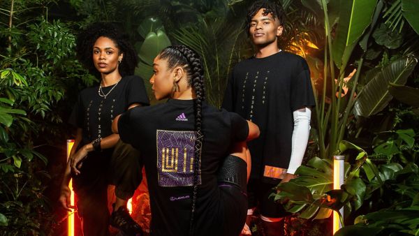 Drei Models posieren vor einer tropischen Kulisse und präsentieren schwarze T-Shirts mit einzigartigen Designs. Das Modell in der Mitte steht mit dem Rücken zur Kamera und zeigt das Adidas-Logo und gemusterte Kunstwerke.