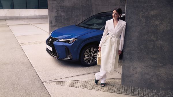 Eine elegante Frau in einem weißen Mantel lehnt an einer Wand neben der ein blauer Lexus UX geparkt ist