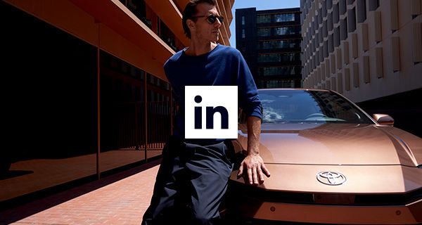 Férfi támaszkodik egy barna Toyota autó motorháztetőjére, háttérben városi épületek és egy LinkedIn logó.