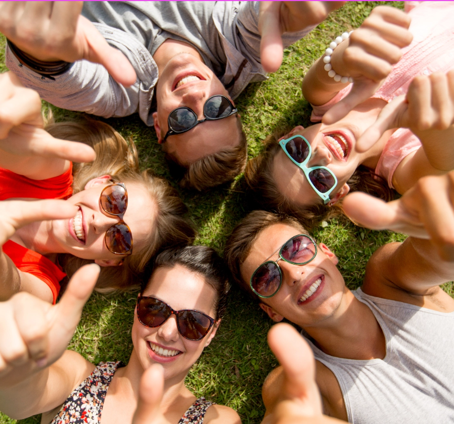 Pięć młodych osób w okularach przeciwsłonecznych leży na trawie na plecach i pokazuje kciuki w górę.