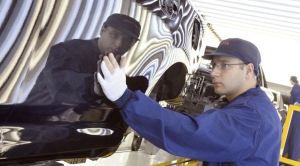 Ein Arbeiter in blauer Uniform inspiziert die glänzende Oberfläche eines schwarzen Autos und konzentriert sich dabei auf die Lackierung vor einem reflektierenden Hintergrund.