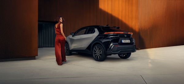 Eine lächelnde Frau geht auf einen geparkten, schwarzen Toyota C-HR zu, der vor einer orangenen Wand steht.
