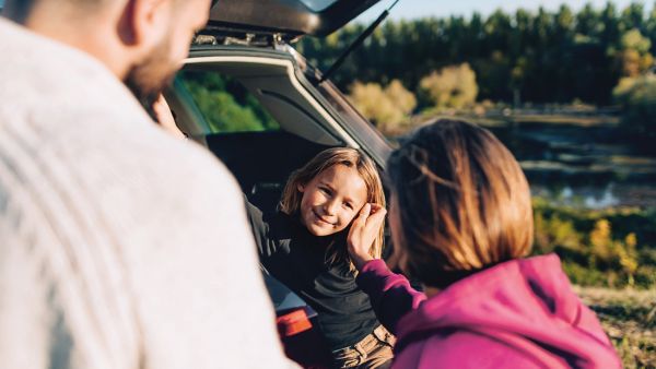 Ein junges Mädchen sitzt auf dem Rücksitz eines offenen Autos und lächelt, während sich zwei Erwachsene aufmerksam mit ihr im Grünen unterhalten.