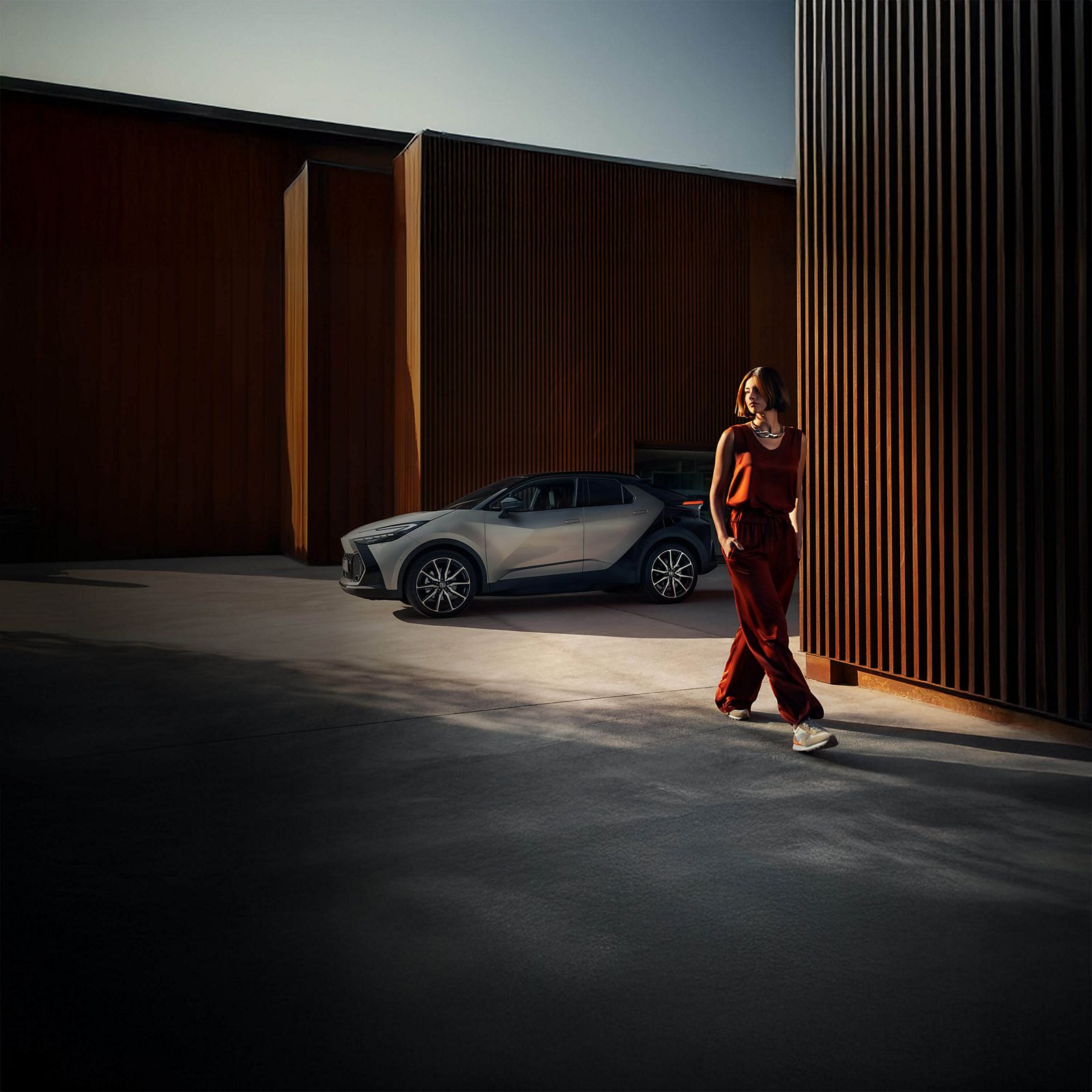 Eine Frau in einem kastanienbraunen Outfit geht durch eine moderne, industrielle Umgebung im Freien mit einem silbernes Auto, das in der Nähe geparkt ist.