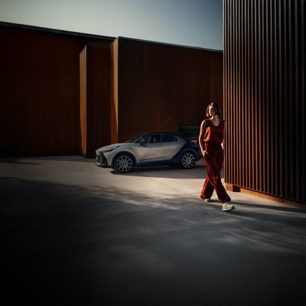 Eine Frau in einem kastanienbraunen Outfit geht durch eine moderne, industrielle Umgebung im Freien mit einem silbernes Auto, das in der Nähe geparkt ist