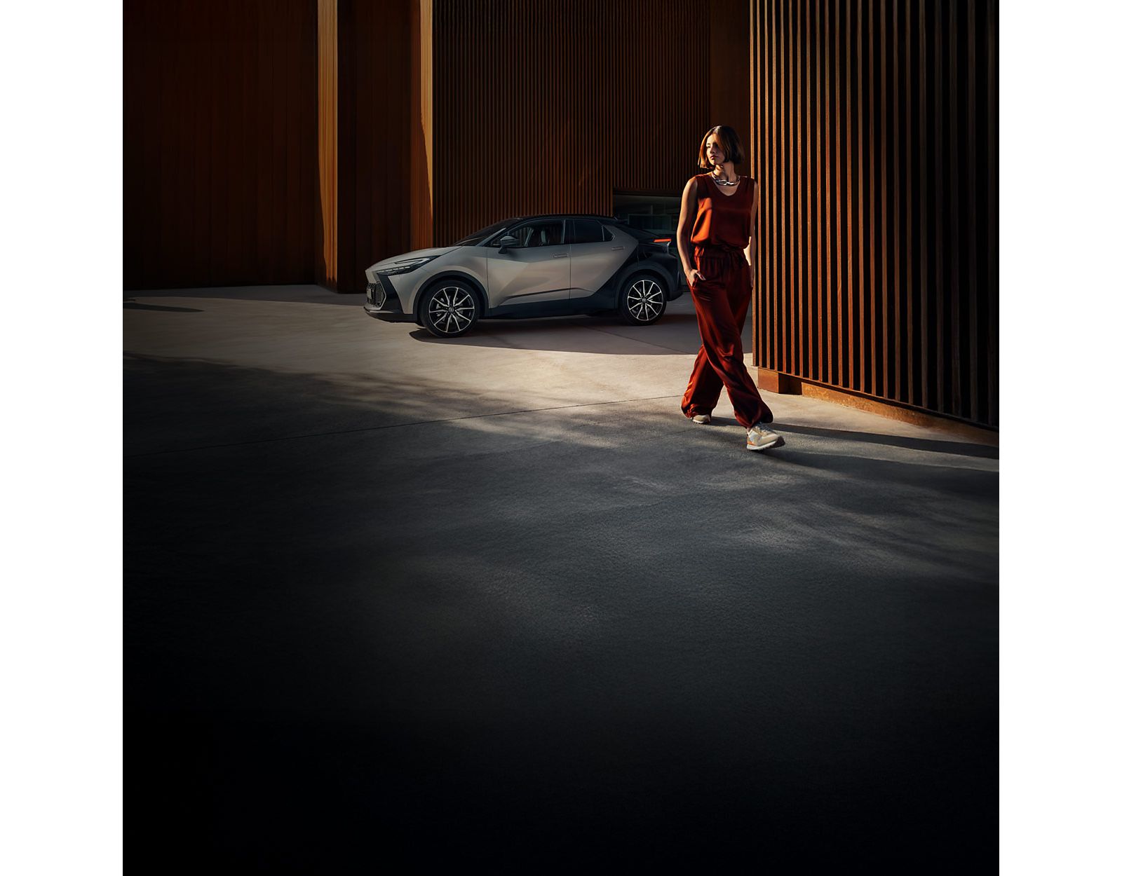 Eine Frau in einem kastanienbraunen Outfit geht durch eine moderne, industrielle Umgebung im Freien mit einem silbernes Auto, das in der Nähe geparkt ist.