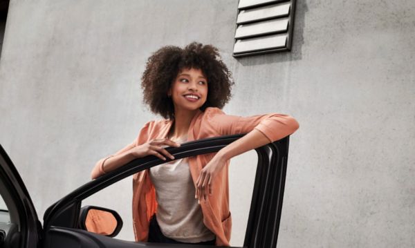 Eine junge Frau legt ihre Arme auf der geöffneten Beifahrertür eines Fahrzeugs ab und schaut lächelnd vom Fahrzeug weg.