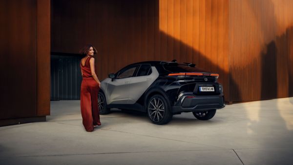 Eine Frau im roten Outfit läuft auf einen grau schwarzen Toyota C-HR zu während im Hintergrund sind die Wände rostbraune Stahlcontainer zu sehen