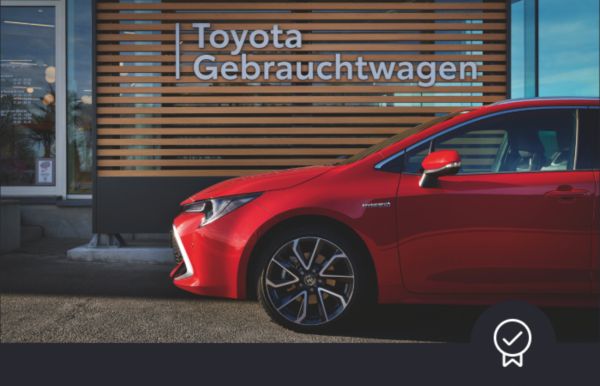 Ein roter Toyota parkt vor einer Holzwand mit der Aufschrift „Toyota Gebrauchtwagen“, die auf ein Gebrauchtwagen-Autohaus hinweist.