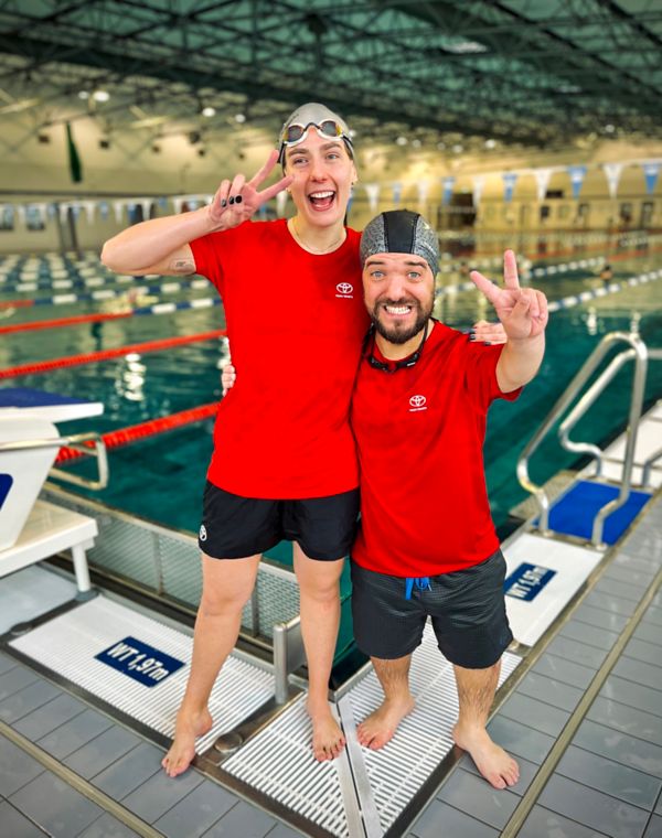 Zwei lächelnde Schwimmer in roten T-Shirts posieren zusammen in einem Schwimmbad, wobei einer ein Peace-Zeichen macht und der andere den Daumen nach oben zeigt