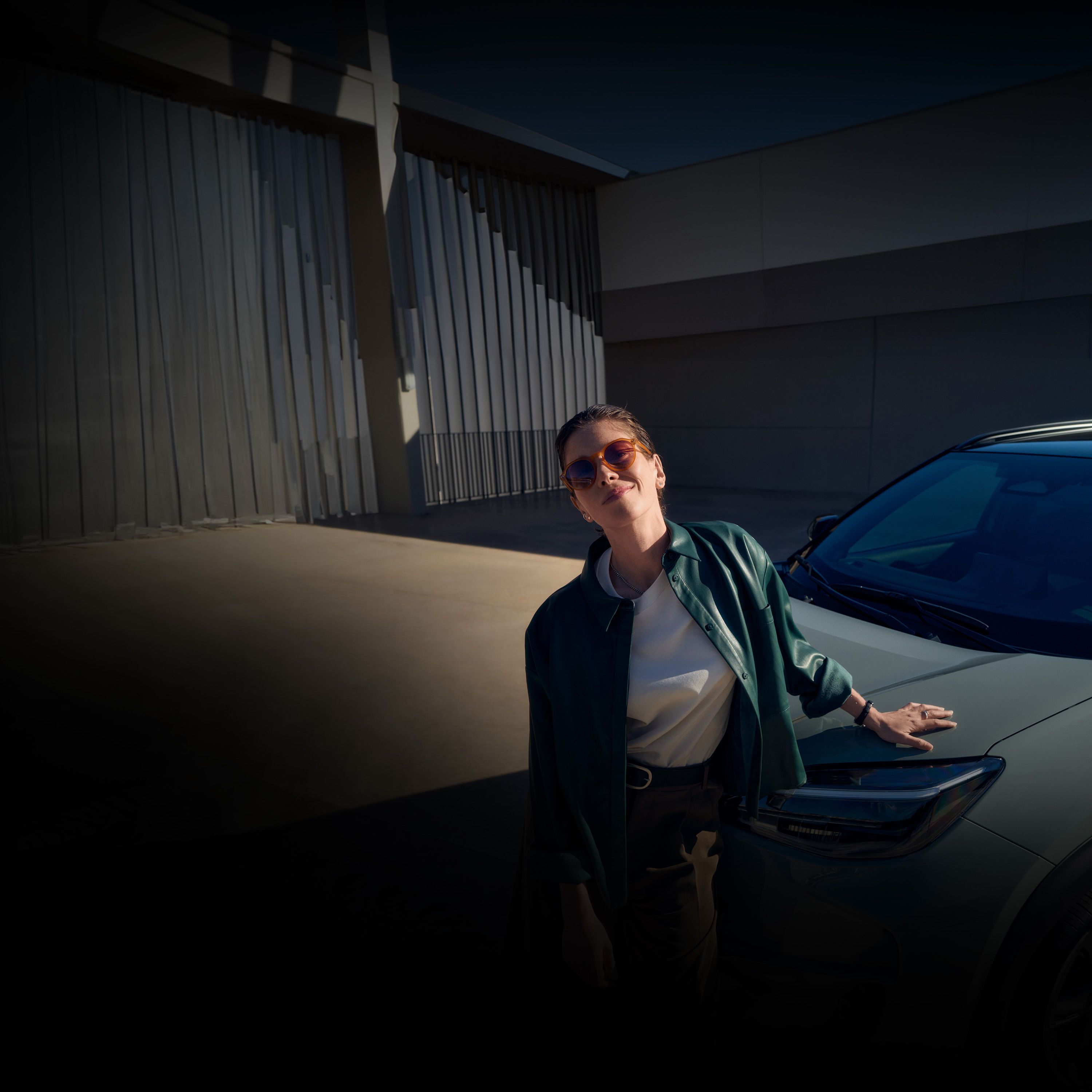 Eine Frau mit Sonnenbrille lehnt sich lächelnd an eine Motorhaube.