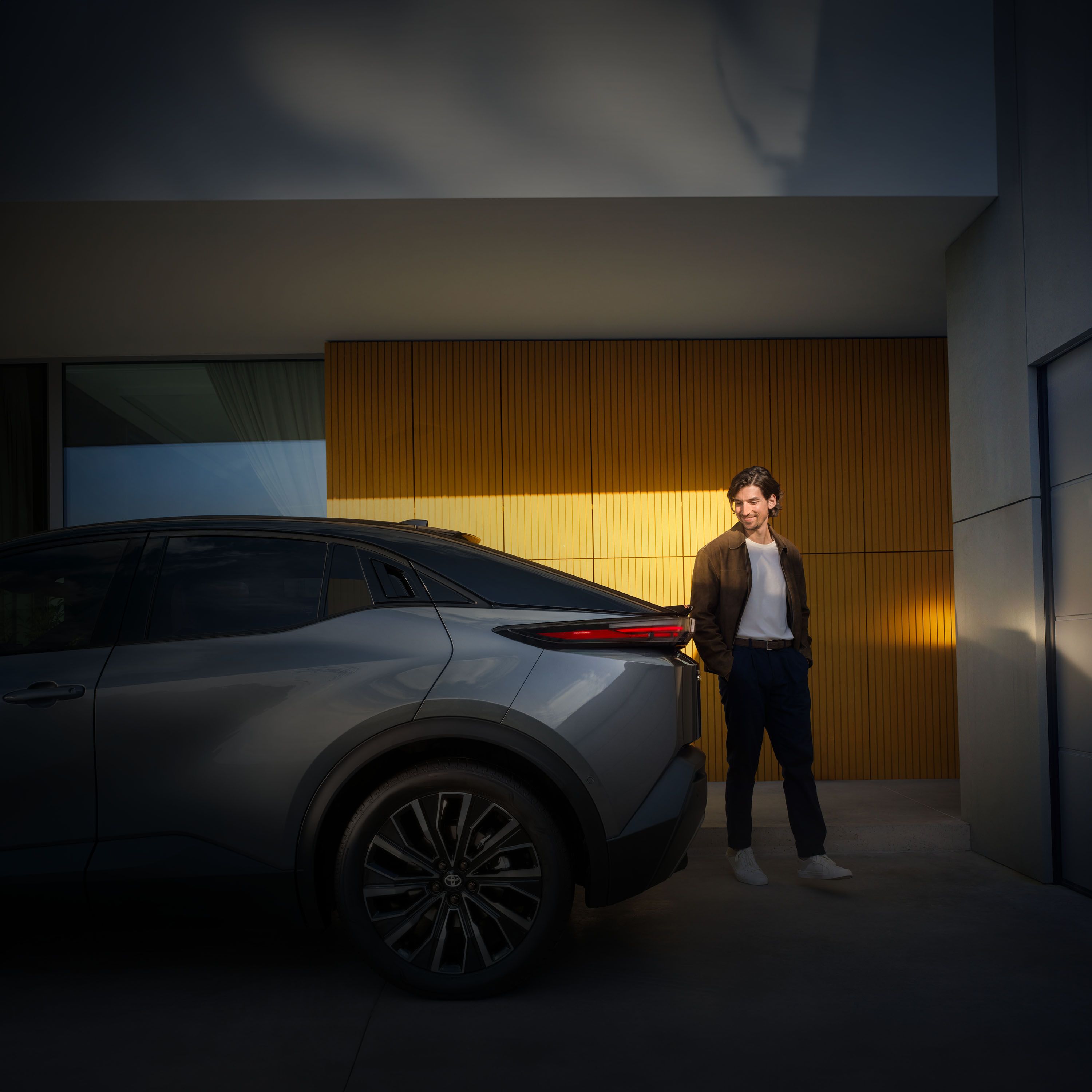 Ein Mann steht lächelnd neben dem Heck eines modernen Autos vor einer gelb beleuchteten Wand; links im Bild sind teilweise Werbetexte und ein Button sichtbar.