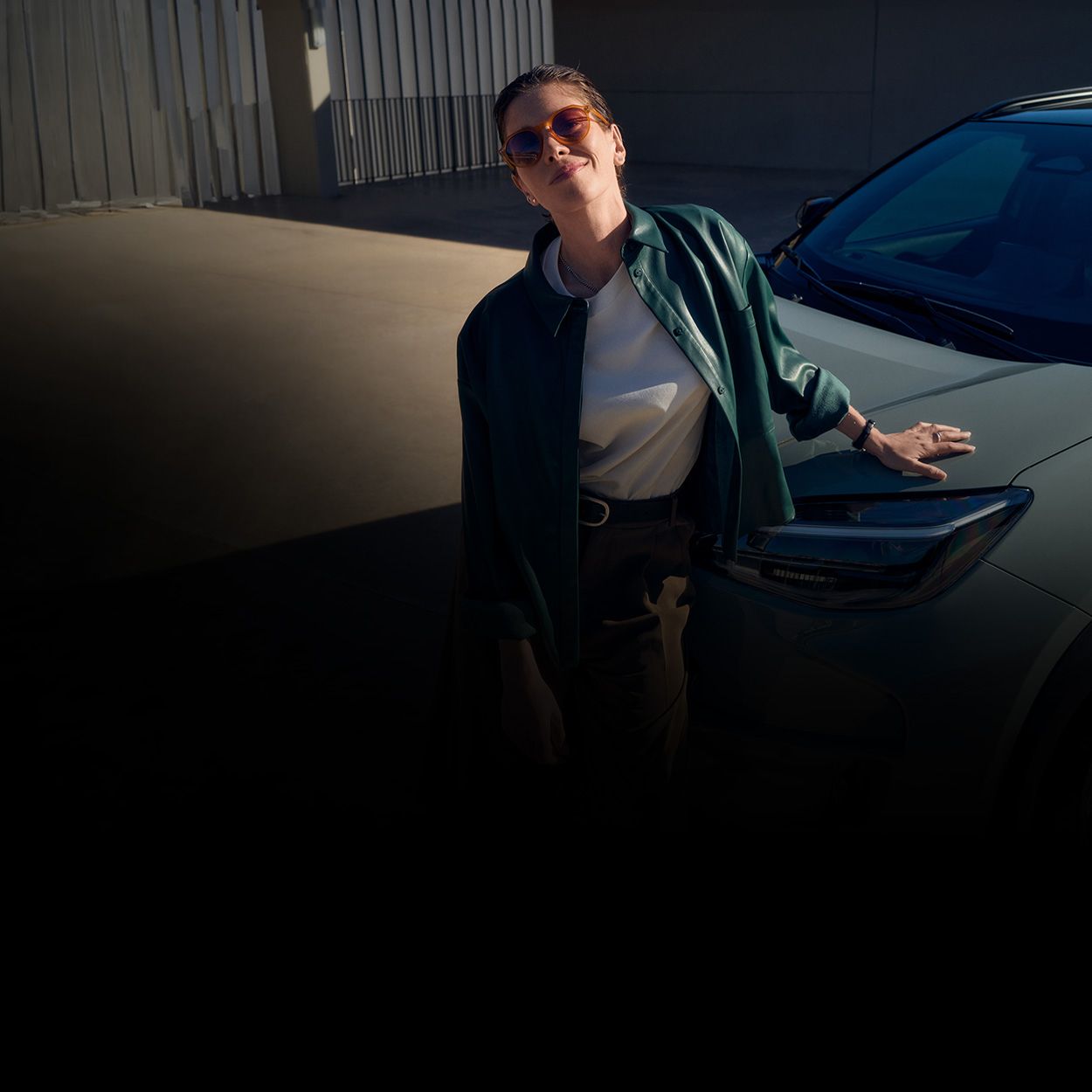 Eine Frau mit Sonnenbrille lehnt sich lächelnd an eine Motorhaube.