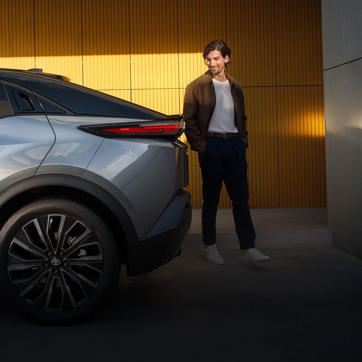 Ein Mann steht lächelnd neben dem Heck eines modernen Autos vor einer gelb beleuchteten Wand; links im Bild sind teilweise Werbetexte und ein Button sichtbar.