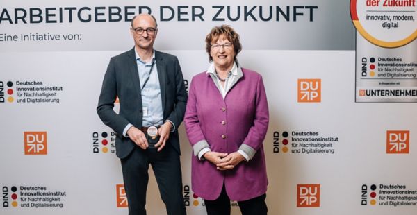 Eine Frau und ein Mann stehen lächelnd nebeneinander vor einem Banner mit der Aufschrift „Arbeitgeber der Zukunft“ und verschiedenen Logos