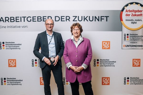 Eine Frau und ein Mann stehen lächelnd nebeneinander vor einem Banner mit der Aufschrift „Arbeitgeber der Zukunft“ und verschiedenen Logos