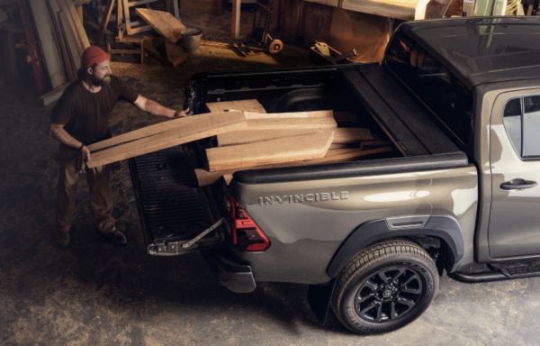 Ein Arbeiter entlädt Holzbretter von der Ladefläche eines grauen Pickup-Trucks in einer Werkstatt, umgeben von Holzbearbeitungswerkzeugen.