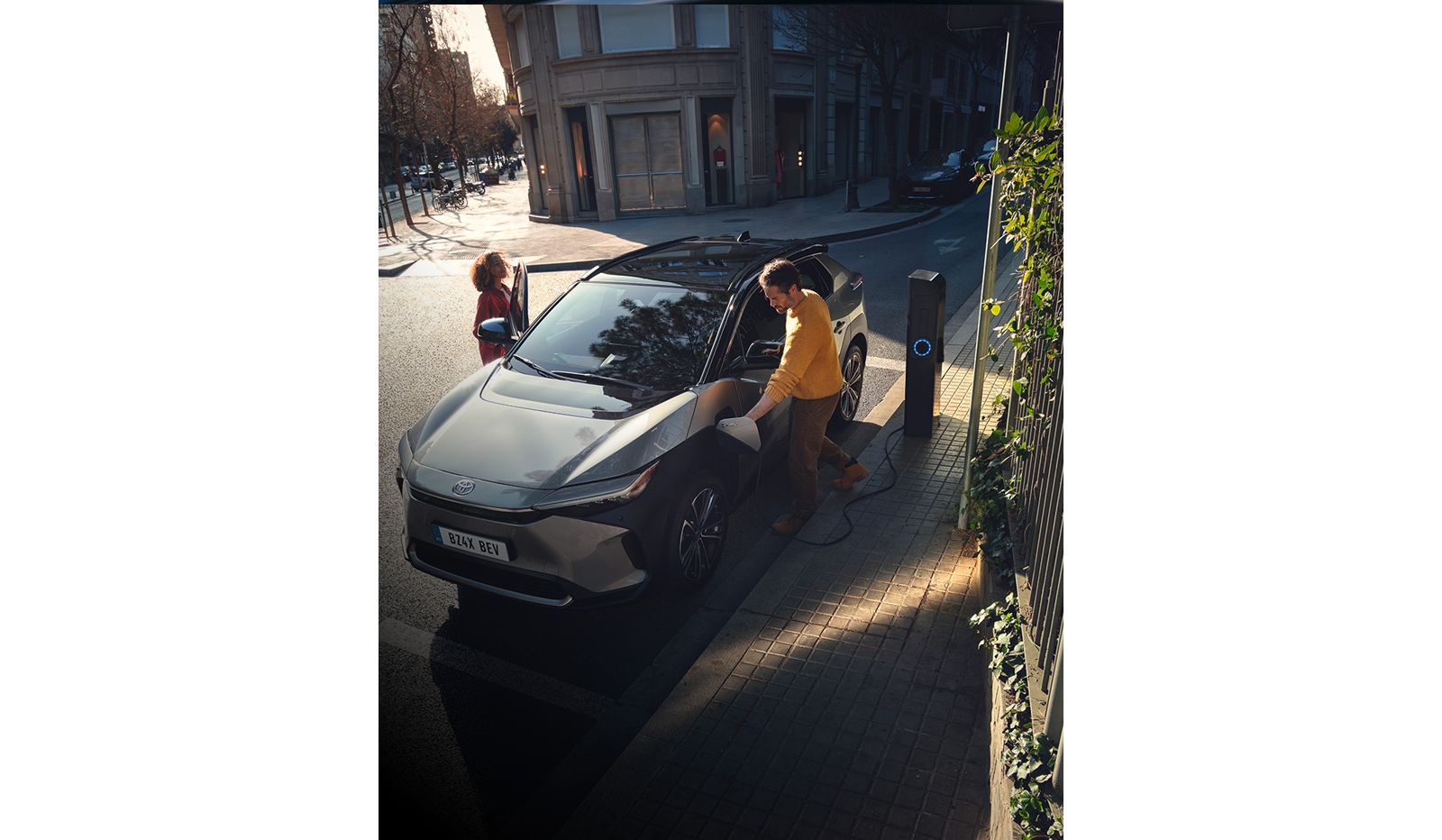 Ein Mann zieht an einer Ladesäule in der Stadt den Ladestecker aus dem geparkten Toyota bZ4X, eine Frau steht an der geöffneten Beifahrertür