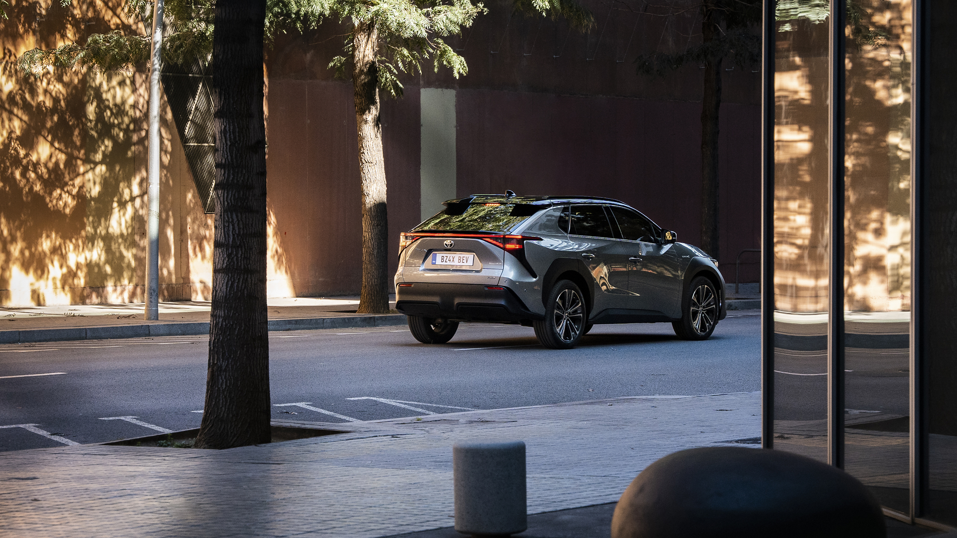 Ein silbergrauer Toyota bZ4X fährt auf einer leeren Straße durch die Stadt, im Vordergrund eine Fensterfront, ein Baum und verschiedene Beton-Elemente