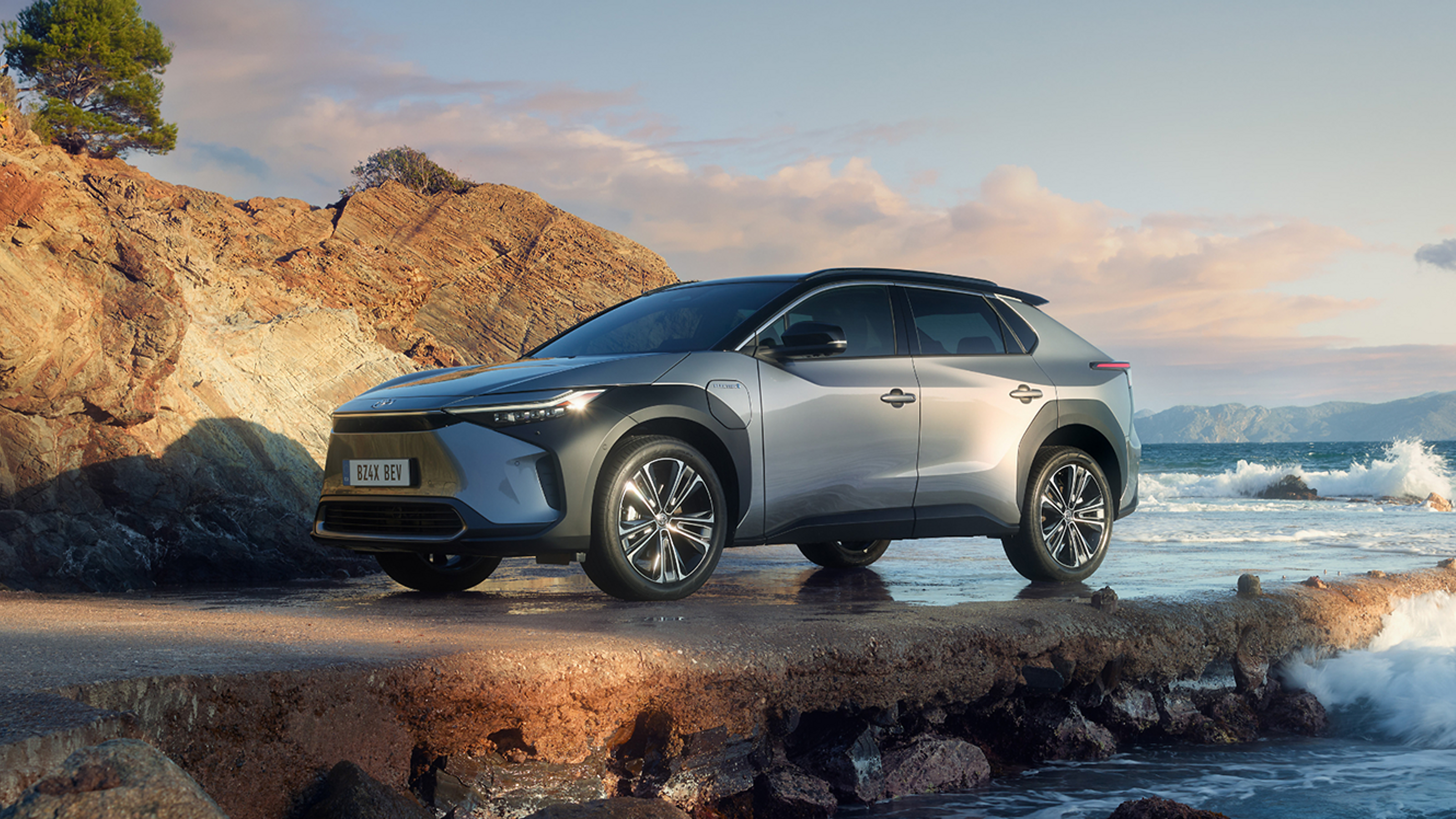 Ein silbergrauer Toyota bZ4X steht an einer Felsenküste, an der sich die Ozeanwellen brechen.