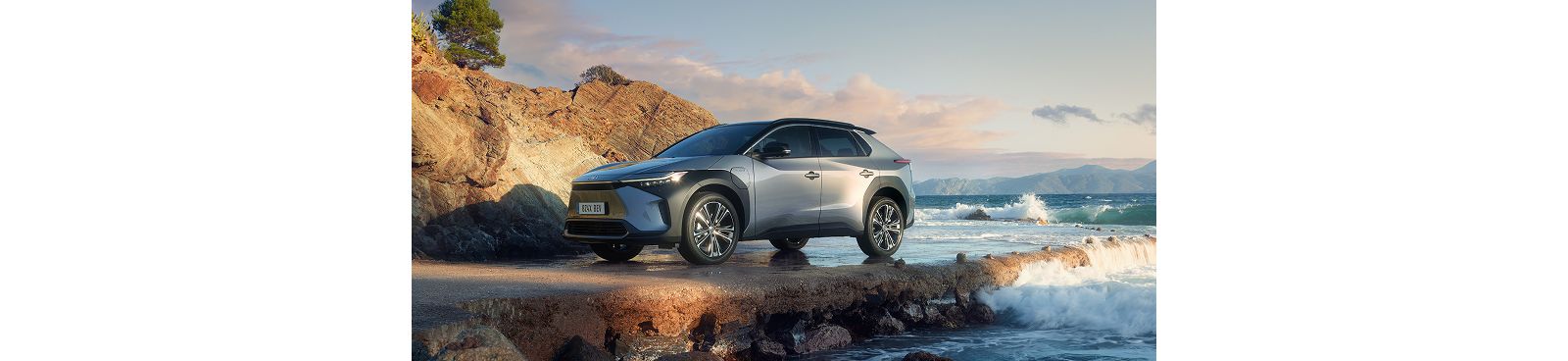 Ein silbergrauer Toyota bZ4X steht an einer Felsenküste, an der sich die Ozeanwellen brechen.
