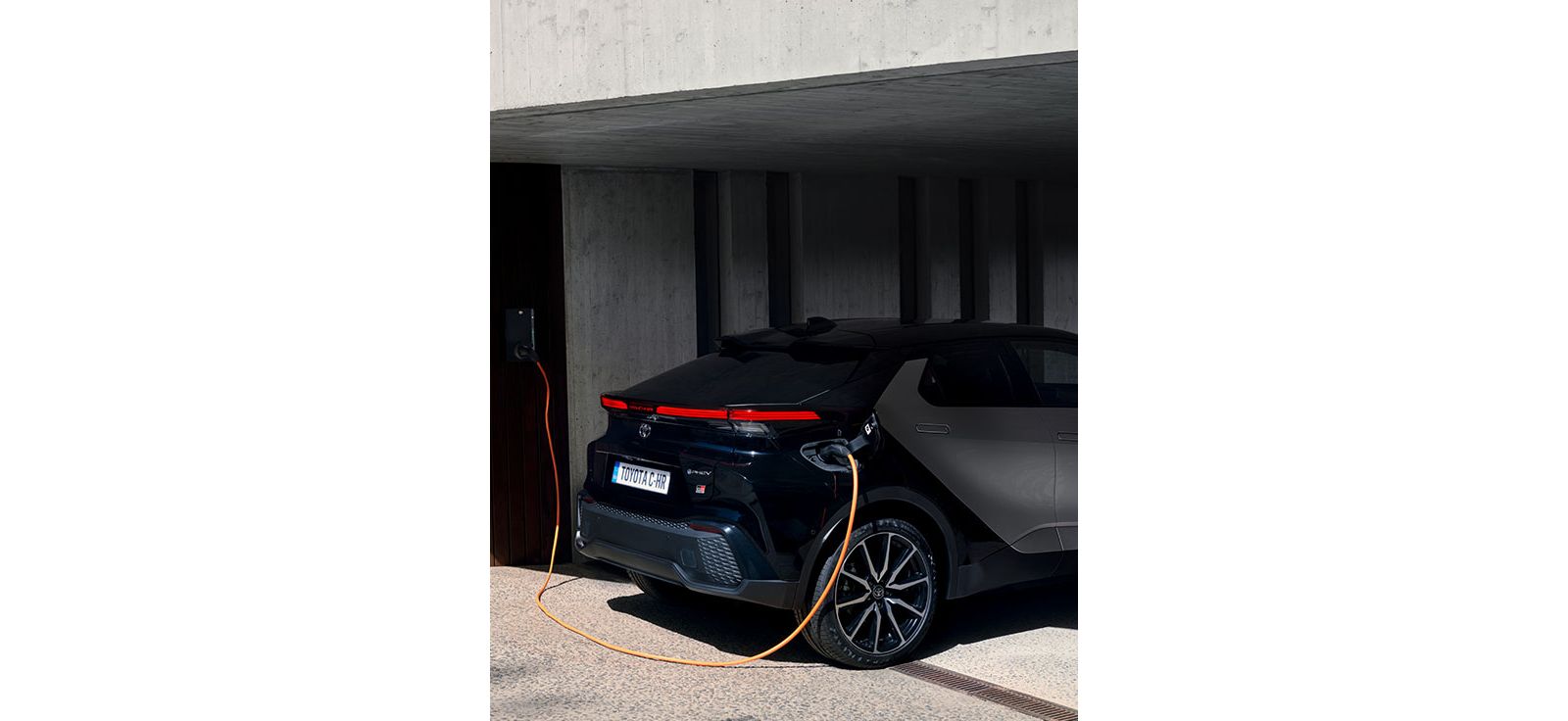 Ein grau-schwarzer Toyota C-HR steht in einem Carport und wird mit einem orangenen Ladekabel geladen