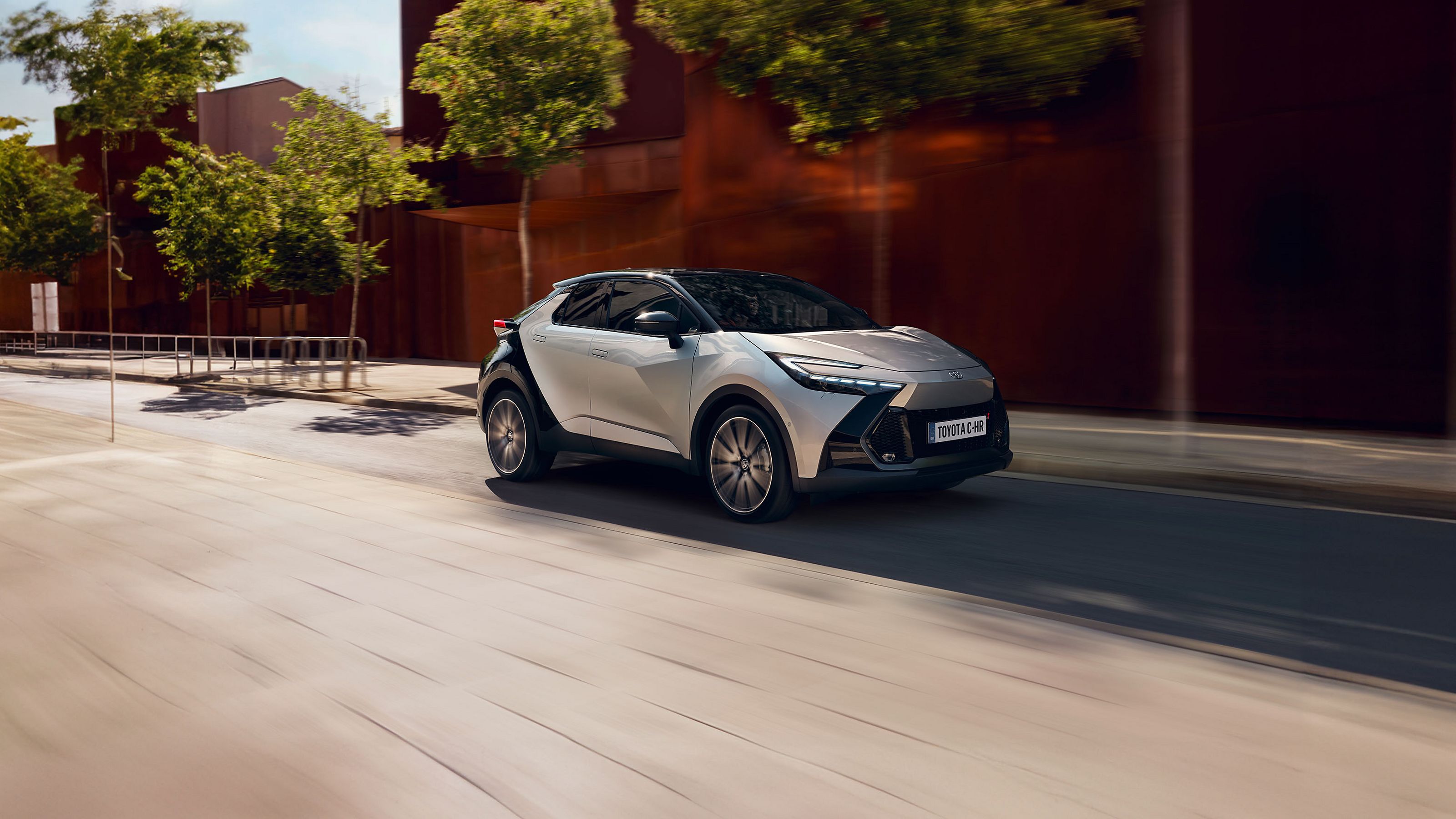 Toyota C-HR in silber fahrend auf einer Straße vor einer roten Wand. Im Hintergrund Bäume. 