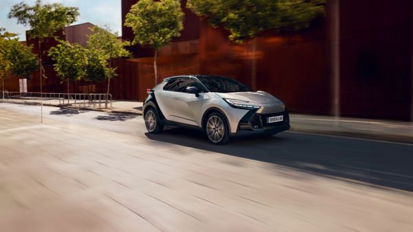 Toyota C-HR in silber fahrend auf einer Straße vor einer roten Wand. Im Hintergrund Bäume. 