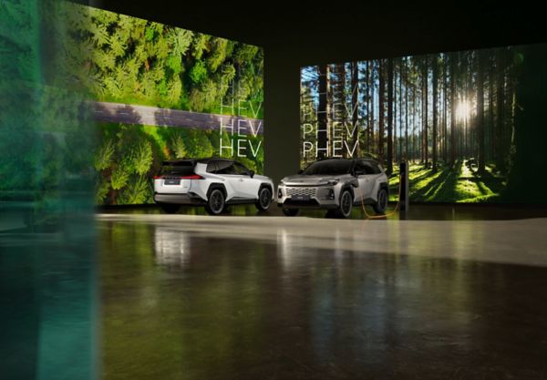 Zwei Toyota SUVs stehen nebeinander in einer Halle, eins davon lädt an einer Ladesäule. Im Hintergrund sind Bildschirme mit dem Text "HEV" hinter dem Linken und "PHEV" hinter dem rechten Fahrzeug.