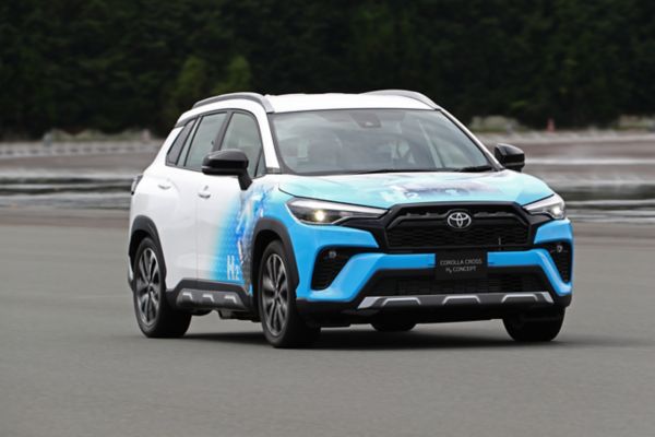 Ein weißer Corolla Cross, vorne blau lackiert und bedruckt mit Wasserstoff-Symbolen von schräg vorne bei der Fahrt auf einer Teststrecke fotografiert