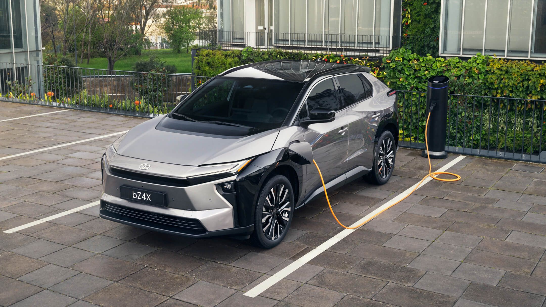 Silberner Elektro-SUV steht auf einem Parkplatz an einer Ladesäule. Im Hintergrund ist ein begrüntes Büro-Gebäude zu sehen.