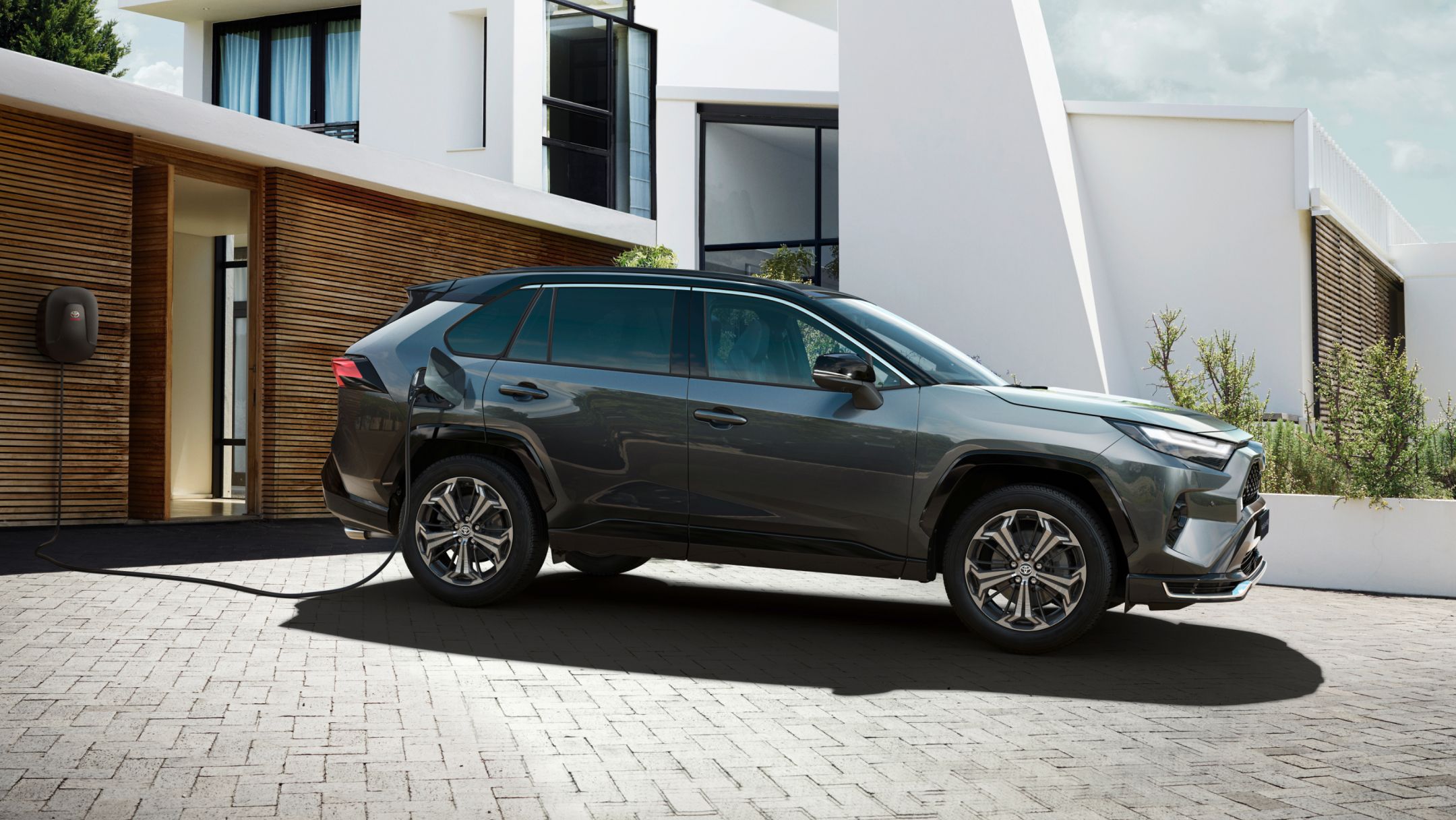 Silberner SUV mit Plug-in-Hybrid-Technologie steht diagonal zur Kamera in einer Studioumgebung und lädt an einer Ladesäule. Im Hintergrund ist auf einem Bildschirm ein Schriftzug mit 'PHEV' zu sehen.