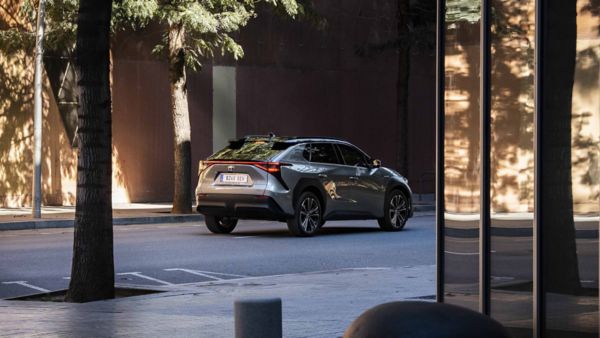 Ein silbergrauer Toyota bZ4X fährt auf einer leeren Straße durch die Stadt, im Vordergrund eine Fensterfront, ein Baum und verschiedene Beton-Elemente