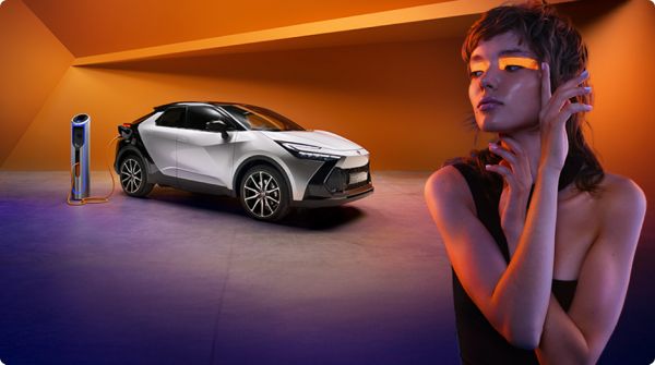 Person mit einem auffälligem orangenen Lidschatten steht vor einem silber-schwarzen Toyota C-HR, der an einer Ladesäule hängt. Im Hintergrund ist eine künstlerisch verformte, orangene Wand zu sehen.