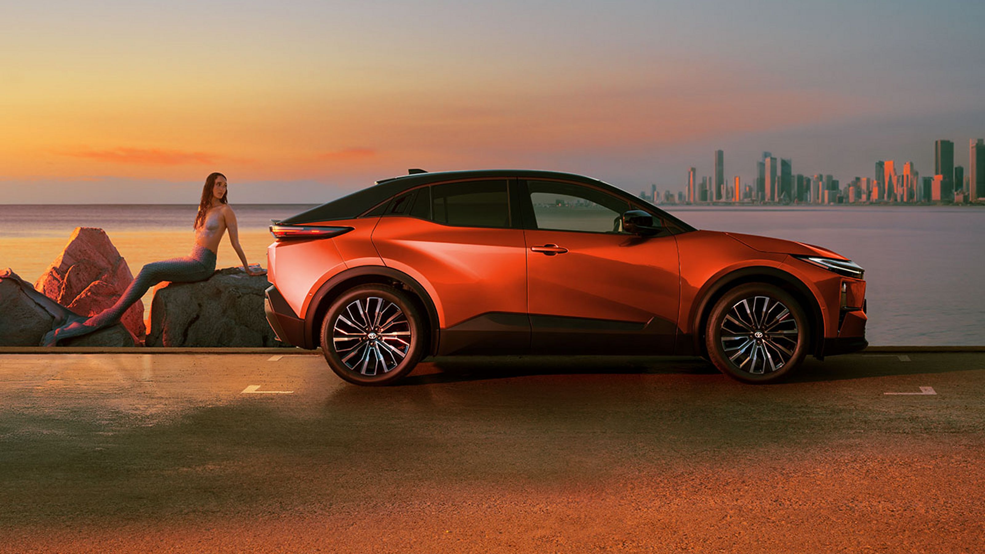 Ein orangefarbener C-HR+ steht bei Sonnenuntergang an einer Küstenstraße; eine Person sitzt entspannt auf Felsen daneben, im Hintergrund sind Meer und eine Skyline zu sehen.