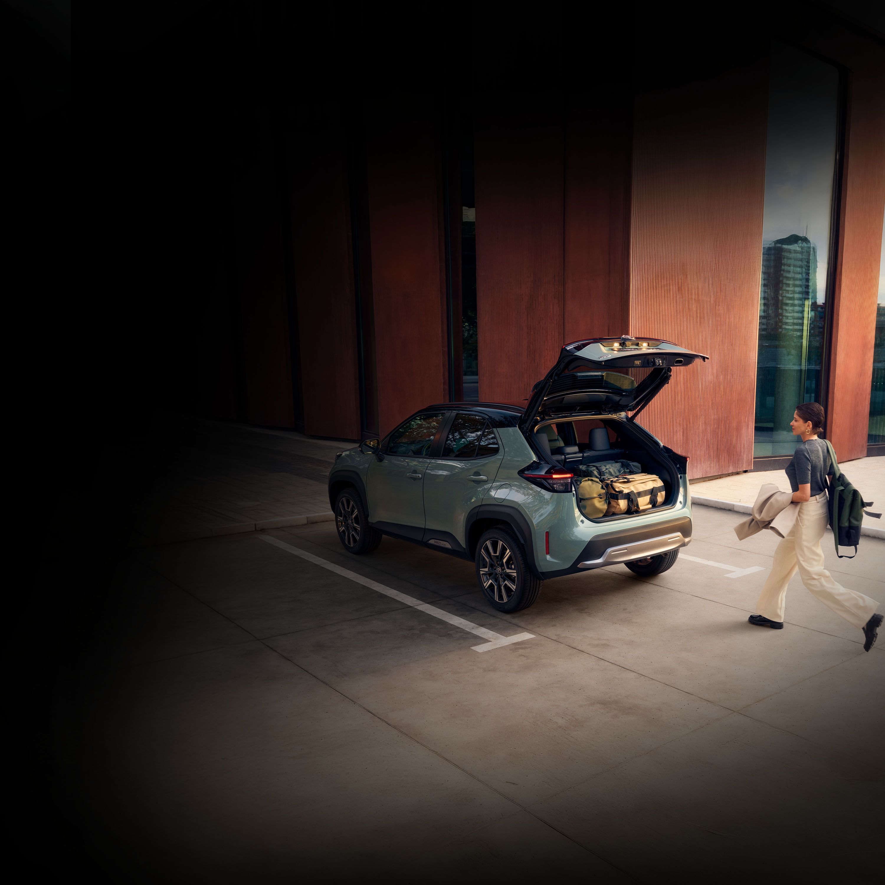 Eine Frau geht auf einen Toyota zu, der geparkt mit geöffneter Kofferraumklappe vor ihr steht. Im Kofferraum befinden sich mehrere Taschen.