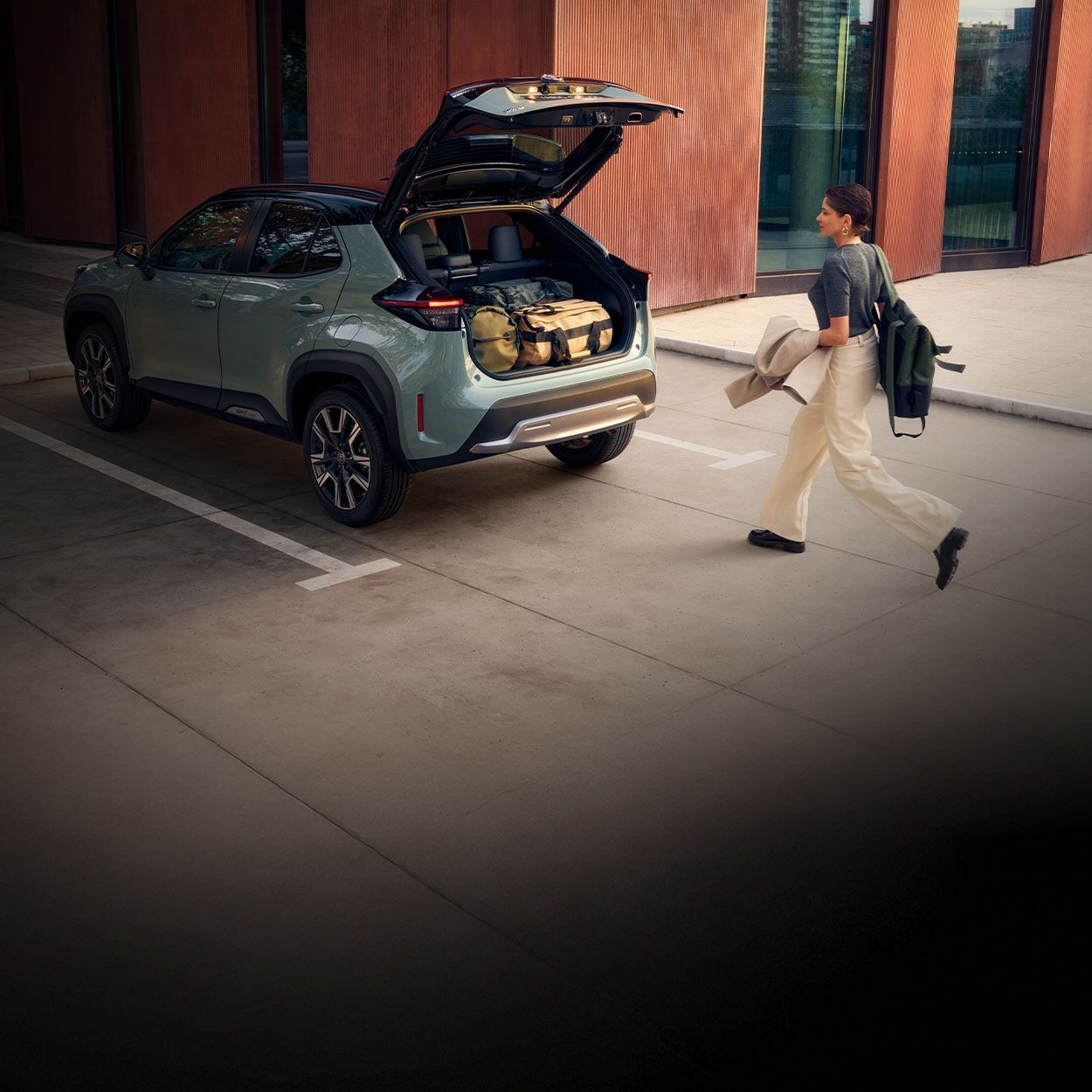 Eine Frau geht auf einen Toyota zu, der geparkt mit geöffneter Kofferraumklappe vor ihr steht. Im Kofferraum befinden sich mehrere Taschen.