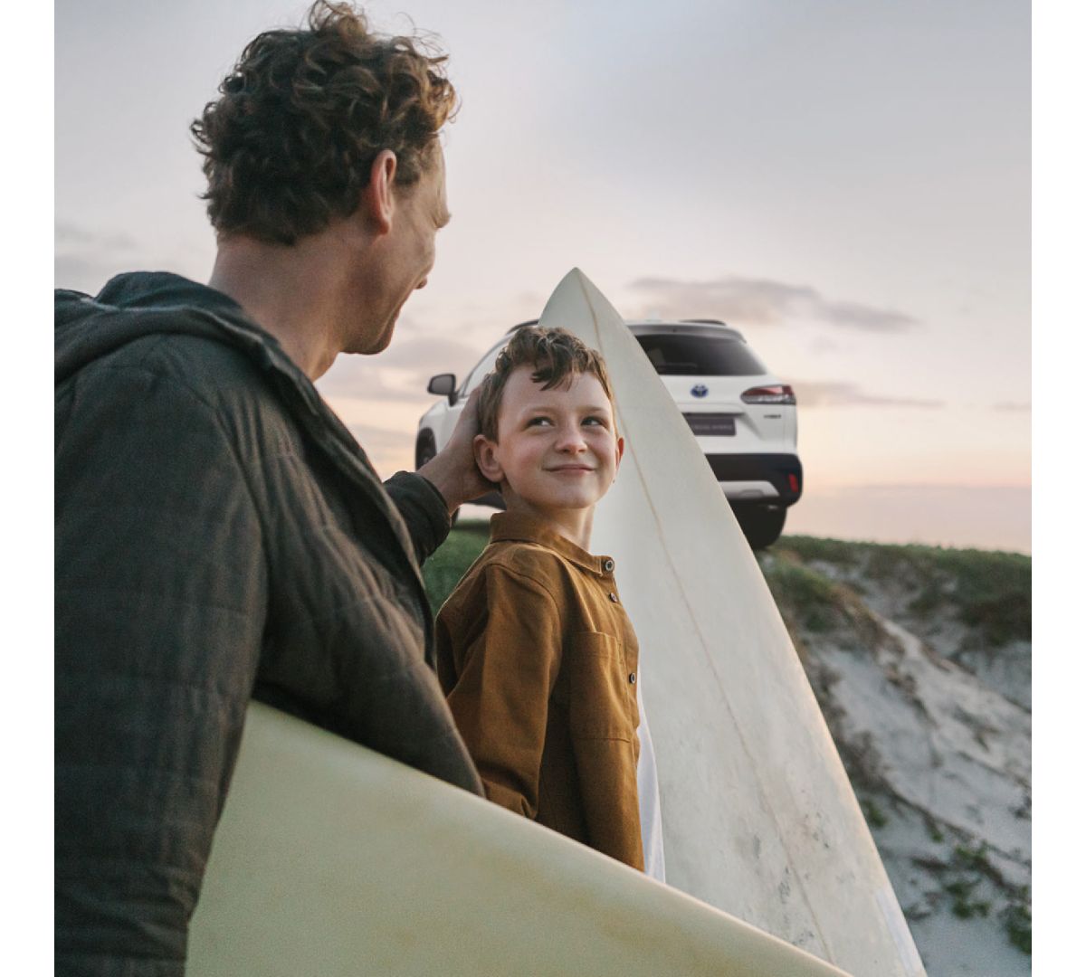Ein Mann sitzt mit einem Kind und ihren Surfbrettern am Strand. Im Hintergrund steht ein geparkter Toyota