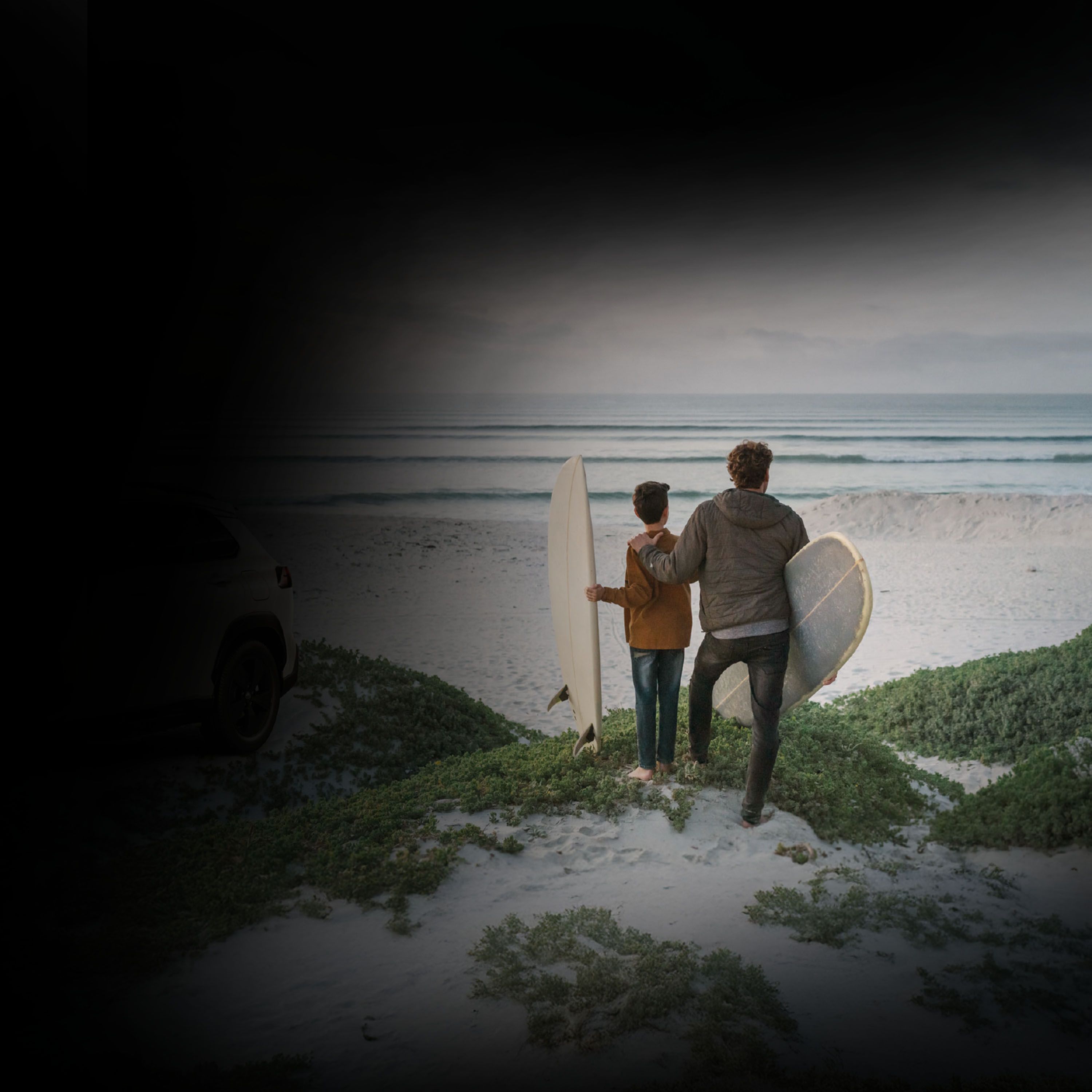 Ein Mann legt einem Kind die Hand auf die Schulter und beide blicken auf einen Strand mit einem Surfbrett unter den Armen