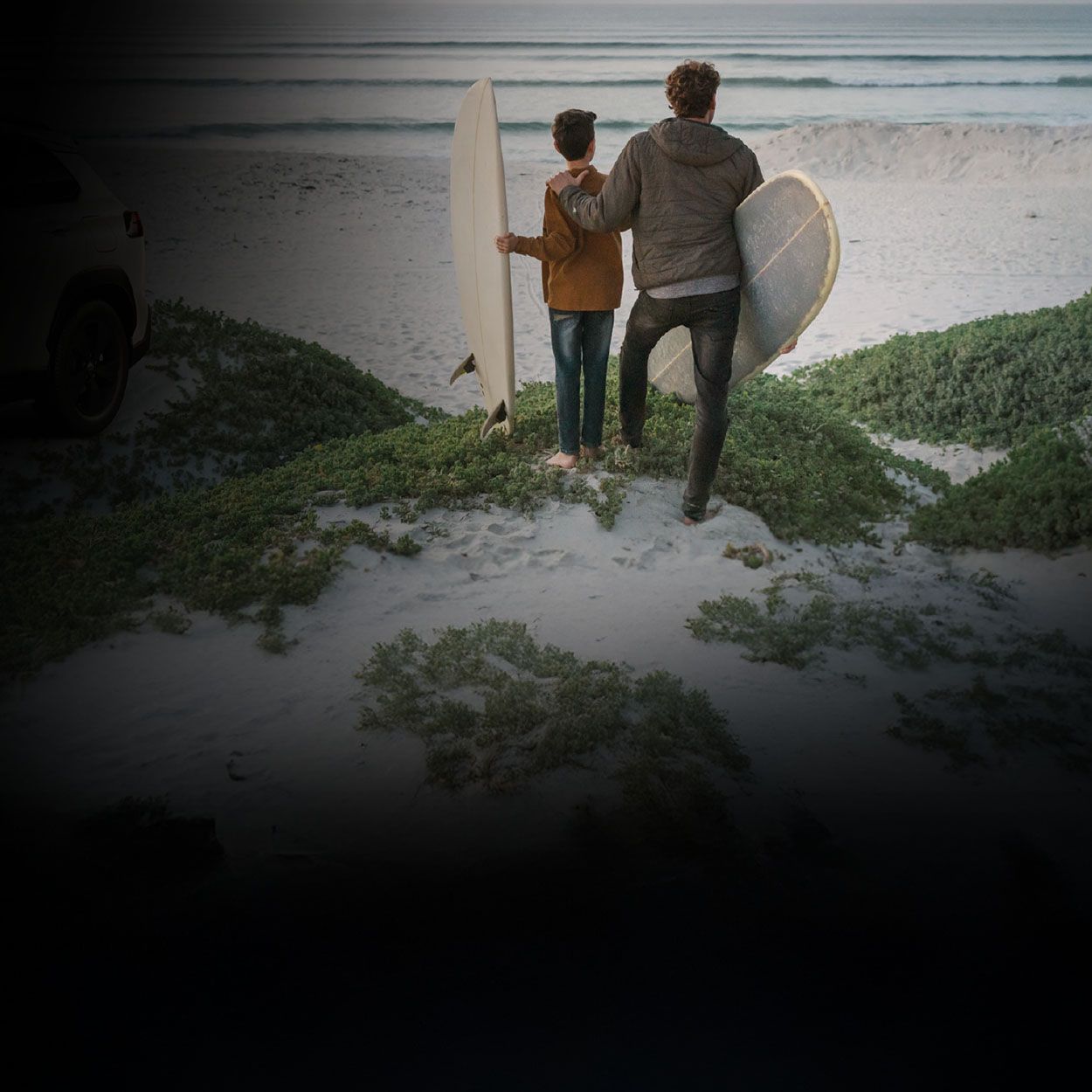 Ein Mann legt einem Kind die Hand auf die Schulter und beide blicken auf einen Strand mit einem Surfbrett unter den Armen