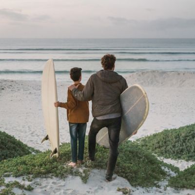 Ein Mann und ein Junge blicken auf einen Strand und jeder hält dabei ein Surfbrett im Arm