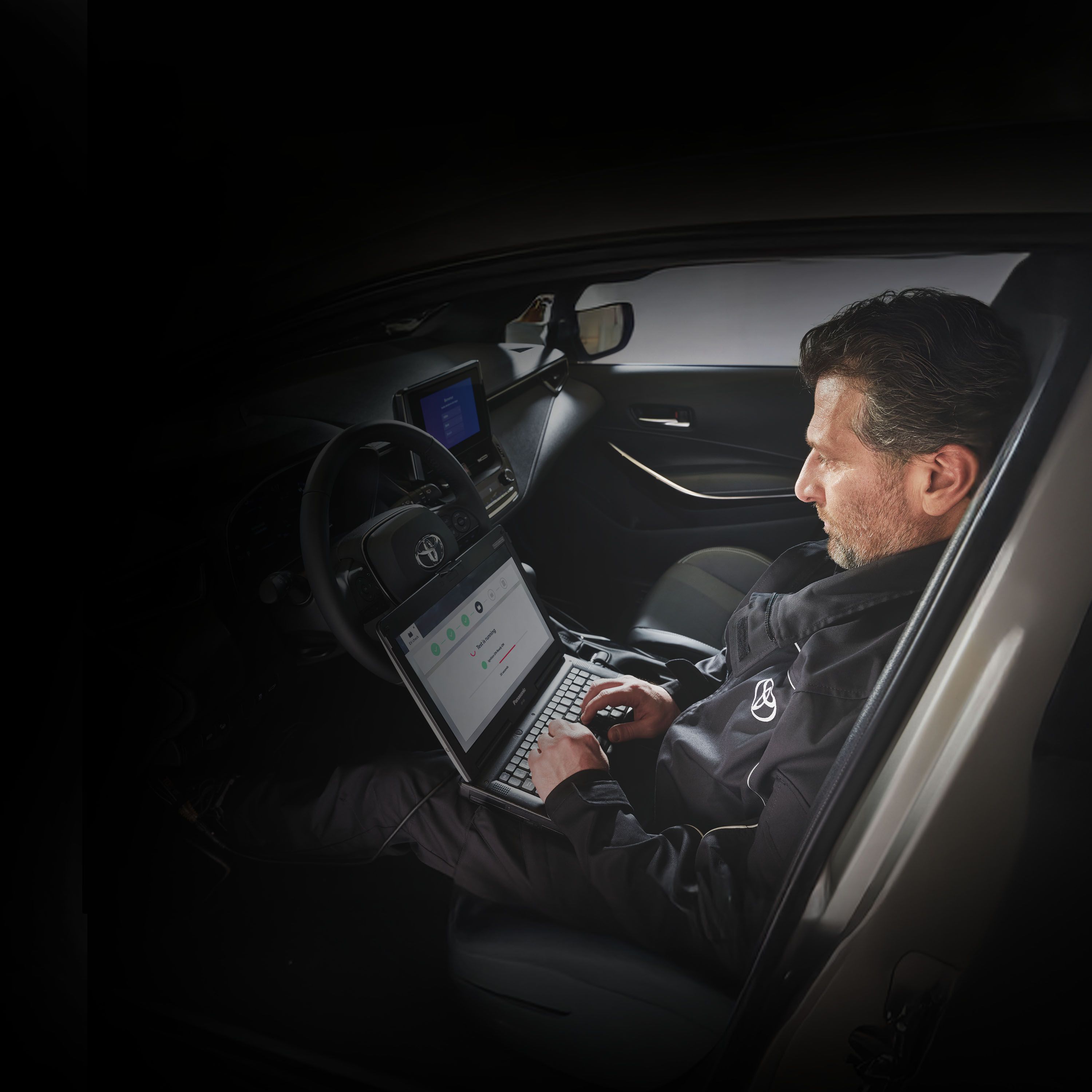 Ein Mechaniker sitzt auf dem Fahrersitz eines Toyota und liest auf seinem Computer, den er auf dem Schoß hat, die Fehlermeldungen des Autos aus.