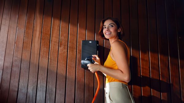 Eine Frau mit orangenem Oberteil hält den Stecker einer schwarzen Wallbox in der Hand, die n einer Holzwand befestigt ist.