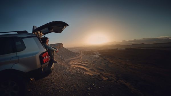 Frau sitzt entspannt im offenen Kofferraum eines SUVs und blickt bei Sonnenuntergang über eine weite Wüstenlandschaft mit kurviger Straße.