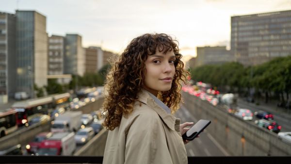 Eine Frau mit einem Handy in der Hand steht auf einer Autobahnbrücke, auf der sich der Verkehr staut, und schaut in die Kamera