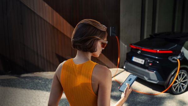 Person hält ein Smartphone in der Hand, auf dem die MyToyota App zu sehen ist. Im Hintergrund steht ein Auto an einer Ladesäule.