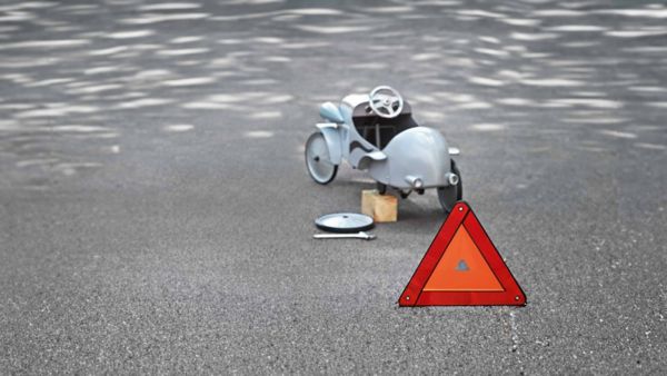 Ein kleines Auto im Vintage-Look mit einem fehlenden Hinterrad steht auf einer Straße neben einem orangefarbenen Warnschild.