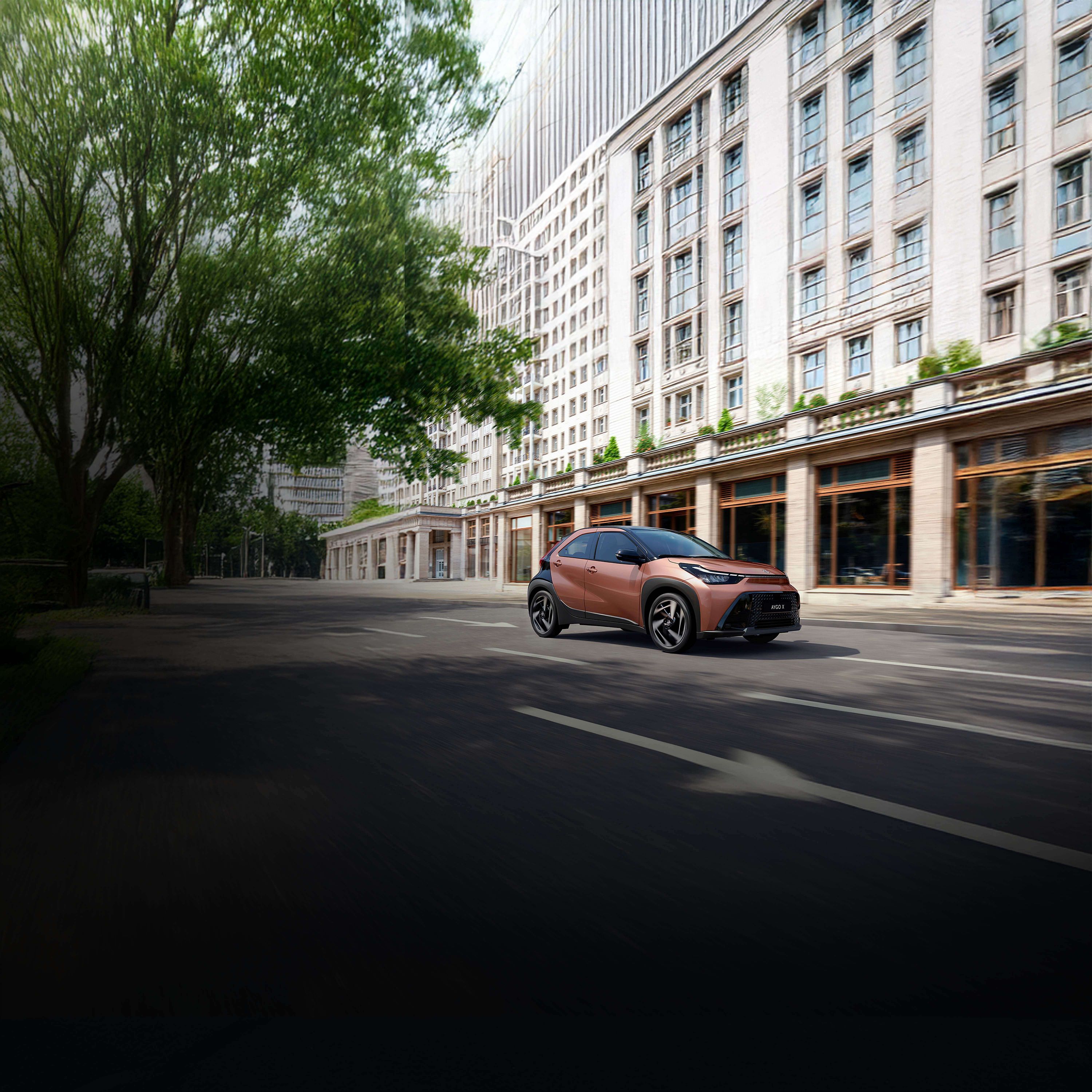 Bronzefarbener Kleinwagen Toyota Aygo X fährt durch eine Innenstadt vor einem Gebäude mit Glasfassade.